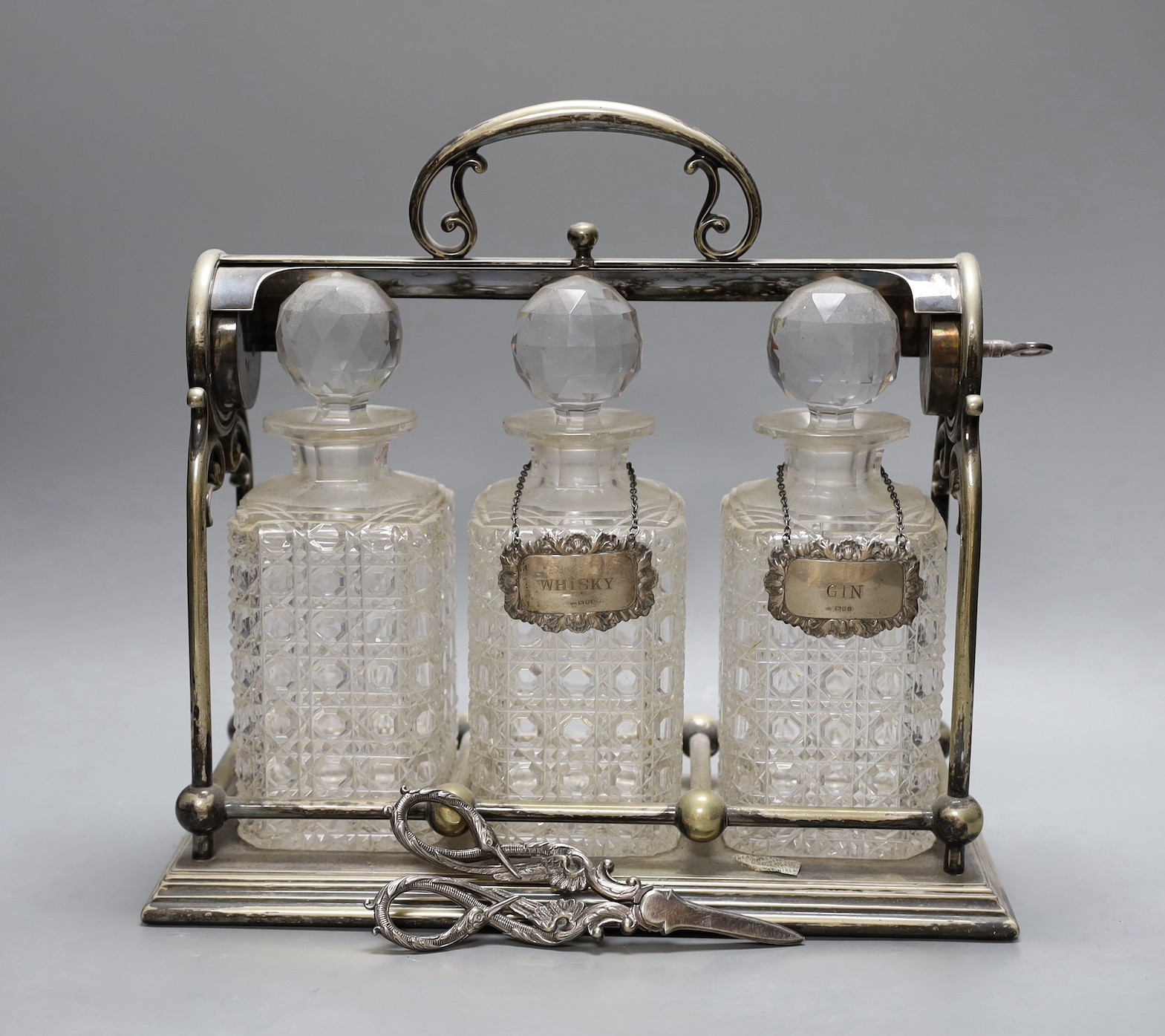 A Victorian electroplate three bottled tantalus, with a silver whisky and gin label plus pair of plated grape scissors, tantalus 33 cms wide